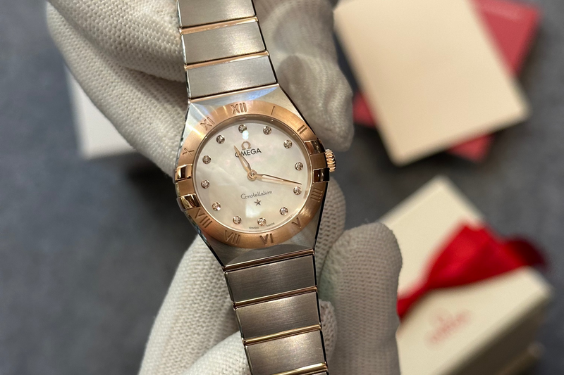 An Omega watch with a two-tone rose gold and stainless steel bracelet, held by a hand wearing a white glove. The watch is shown with its original packaging boxes in the background.