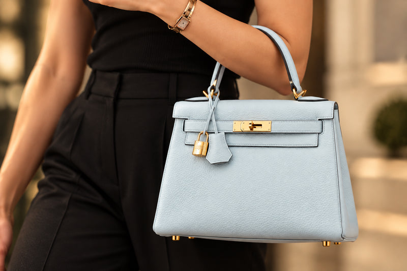 A woman in a black ribbed tank top and black trousers carries a structured light blue leather handbag with gold-tone hardware and a clochette, photographed in an outdoor urban setting.